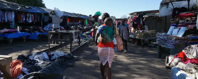 An open-air clothes market on a sunny day: stalls on both sides, and a woman carrying a baby on her back walking between them