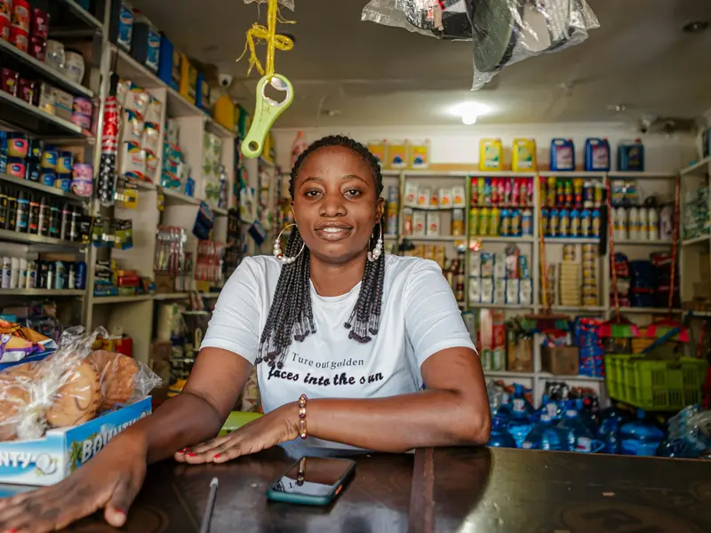 A shopkeeper behind the counter of a small grocery shop, shelves of goods behind her and her phone on the counter